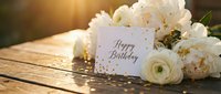 Happy Birthday card with white peonies and ranunculus on rustic wooden table, golden confetti, warm sunlight bokeh background.