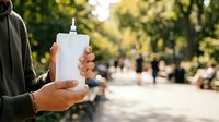 Man holding blank white sunscreen bottle outdoors in park with blurred background, suitable for skincare product marketing and wellness advertising.