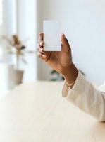 Woman with dark skin tone holding blank white card in bright minimalist interior with potted plant, wearing beige linen sleeve. Mockup for branding, business cards, invitations.