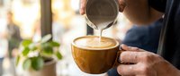 Barista pouring steamed milk into coffee cup creating latte art in cozy cafe setting with warm natural lighting for coffee shop marketing materials.