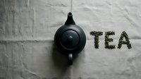 Black ceramic teapot on linen fabric beside word TEA spelled with dried green tea leaves, top view flat lay composition.