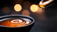 Liquid amber drop falling into dark container creating ripple effect, with soft bokeh lights in background. Macro photography captures honey or syrup motion.