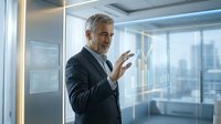Mature Caucasian businessman in dark suit touching transparent digital interface screen in modern glass office with city view background.