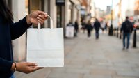 Person holding blank white paper bag on busy urban street with blurred pedestrians and storefronts in background for retail and shopping concepts.