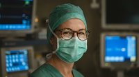 Female surgeon wearing surgical mask and cap with glasses in modern operating room with medical monitors and equipment in background.