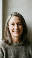 Mature woman with shoulder-length gray hair smiling at camera, wearing neutral knit sweater against white wall backdrop. Portrait suitable for business, healthcare, education and lifestyle editorial use.