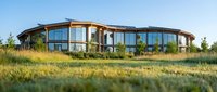 Modern curved residential building with extensive glazing situated in natural grassland landscape during golden hour, showcasing contemporary architecture and sustainable design for real estate and architectural publications.