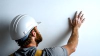 Construction worker in white hard hat inspecting wall surface quality during renovation, wearing gray workwear, side view of professional builder at work.
