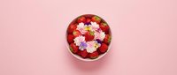 Fresh strawberries and colorful cream flowers in a white bowl on a pastel pink background, top view flat lay for food blog or menu.