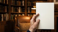 Senior man holding blank white paper sheet in library with leather armchair and bookshelves, suitable for mockups, advertising, and editorial layouts.