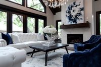 Modern luxury living room with navy velvet chairs, white tufted sofa, crystal chandelier, abstract blue wall art and fireplace. Contemporary interior design with natural light.