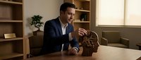 Professional businessman in dark suit examining wooden brain puzzle at office desk, symbolizing problem-solving and strategic thinking in corporate environment.