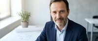 Mature male professional with dark hair and beard wearing navy blazer and light blue shirt, seated at desk with architectural plans and potted plant in modern office environment.