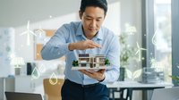 Smiling Asian male architect in blue shirt examining residential house model with sustainable energy icons displayed in modern office environment.