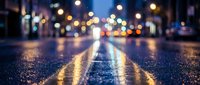 Wet asphalt road surface with yellow lane markings reflecting colorful city lights and bokeh effects during evening hours creating atmospheric urban scene.