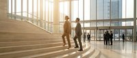 Two professional businessmen in suits having discussion on modern office building stairs with glass windows and natural sunlight streaming through lobby.