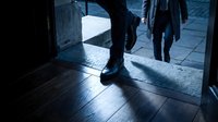 Person wearing black shoes standing in doorway with sunlight streaming across wooden floor, urban street scene visible outside.