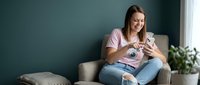 Young Caucasian woman in pink shirt sitting on beige armchair holding small white puppy dog in cozy living room with teal wall and houseplant at home.