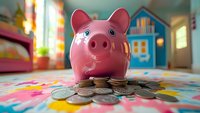 Pink ceramic piggy bank with scattered coins on colorful floral pattern surface against blurred playful background setting, representing savings and financial education.