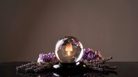 Crystal ball with glowing candle reflection surrounded by amethyst crystals and lavender sprigs on dark reflective surface, mystic spiritual atmosphere.
