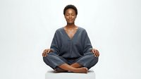 Young African woman in casual blue shirt sitting cross-legged in meditation pose against white background, suitable for wellness, mindfulness and lifestyle design projects.