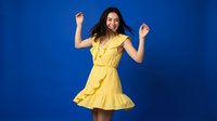 Happy young woman dancing in bright yellow summer dress against vibrant blue studio background expressing joy and positive energy for lifestyle content.