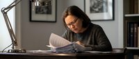Middle-aged Asian woman with glasses reading documents at desk with lamp in modern home office, focused on paperwork and administrative tasks.