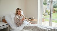 Middle-aged Caucasian woman relaxing in bed with coffee cup, wearing cozy beige robe by large window with garden view for wellness and morning routine concepts.