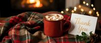 Cozy winter scene with hot cocoa mug, marshmallows, thank you card, plaid blanket, and fireplace bokeh lights in the background.
