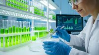 Female scientist in white lab coat and blue gloves conducting research with test tubes containing green liquid in modern laboratory setting.