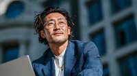 Asian businessman with glasses and messy hair smiling confidently outdoors in blue suit jacket holding laptop computer for corporate success concepts.
