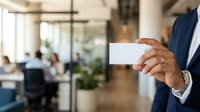 Businessman in navy suit holding blank white business card in modern office environment with colleagues and plants in background, suitable for corporate branding and professional networking concepts.