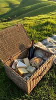 Wicker picnic basket filled with wine, cheese, and bread on rolling green hillside during golden hour sunlight, perfect for outdoor dining and countryside gatherings.