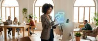Woman with curly dark hair, mixed ethnicity, wearing brown blazer, holding digital tablet with holographic interface in modern office workspace with diverse colleagues and natural light.