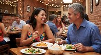 Mature couple enjoying romantic dinner date at cozy brick restaurant with string lights, sharing meal and wine while laughing together in warm atmosphere.