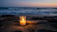 Glowing candle in glass jar on sandy beach at sunset with ocean waves and driftwood creating peaceful evening atmosphere for meditation and relaxation.