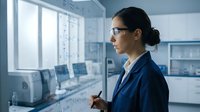 Young Caucasian female scientist in blue lab coat and glasses writing mathematical formulas on a transparent glass board in a modern laboratory.