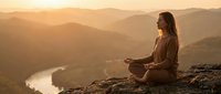Young woman meditating in lotus position on mountain cliff at golden sunset with layered hills and winding river valley below creating peaceful mindfulness scene.