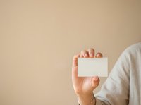 Person holding blank white business card in hand against neutral beige background, suitable for branding, corporate identity, and marketing materials.