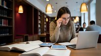 Young woman student studying with laptop and books in modern library with warm lighting. Academic research, education, learning concept for university life.