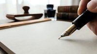 Hand holding fountain pen writing on blank paper with vintage ink bottles and wooden accessories in background for calligraphy and correspondence.