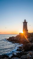 Lighthouse beacon shining bright during golden hour sunset over rocky coastline with ocean waves and seagulls flying in colorful sky for travel concepts.
