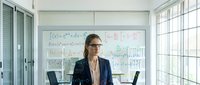 Professional businesswoman with glasses standing in modern office conference room with mathematical equations on whiteboard behind her.