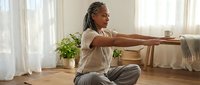 Young African American woman with dreadlocks practicing yoga stretching exercise at home in bright living room with natural light from window.