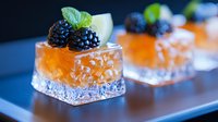Fresh blackberries and cucumber garnish elegant appetizer bites on crystal glass squares filled with bright orange salmon or seafood mousse against dark background.