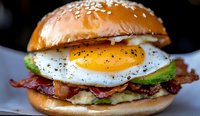 Gourmet breakfast burger with sunny side up egg, crispy bacon strips, fresh avocado slices, and sesame seed bun, served on white plate. Close up food photography.