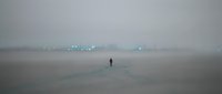 Solitary figure standing on misty beach with distant cargo ships on horizon, moody atmospheric seascape for editorial and conceptual projects.
