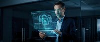 Businessman in dark suit holding transparent digital tablet displaying glowing padlock and security network interface in modern server room environment for cybersecurity concepts.