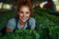 Young female farmer with red curly hair smiling while tending to fresh organic kale crops in sustainable greenhouse garden. Natural lighting highlights genuine joy.