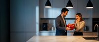 Young professional couple sharing romantic moment with gift box in modern luxury kitchen interior with pendant lighting and sleek cabinetry design.